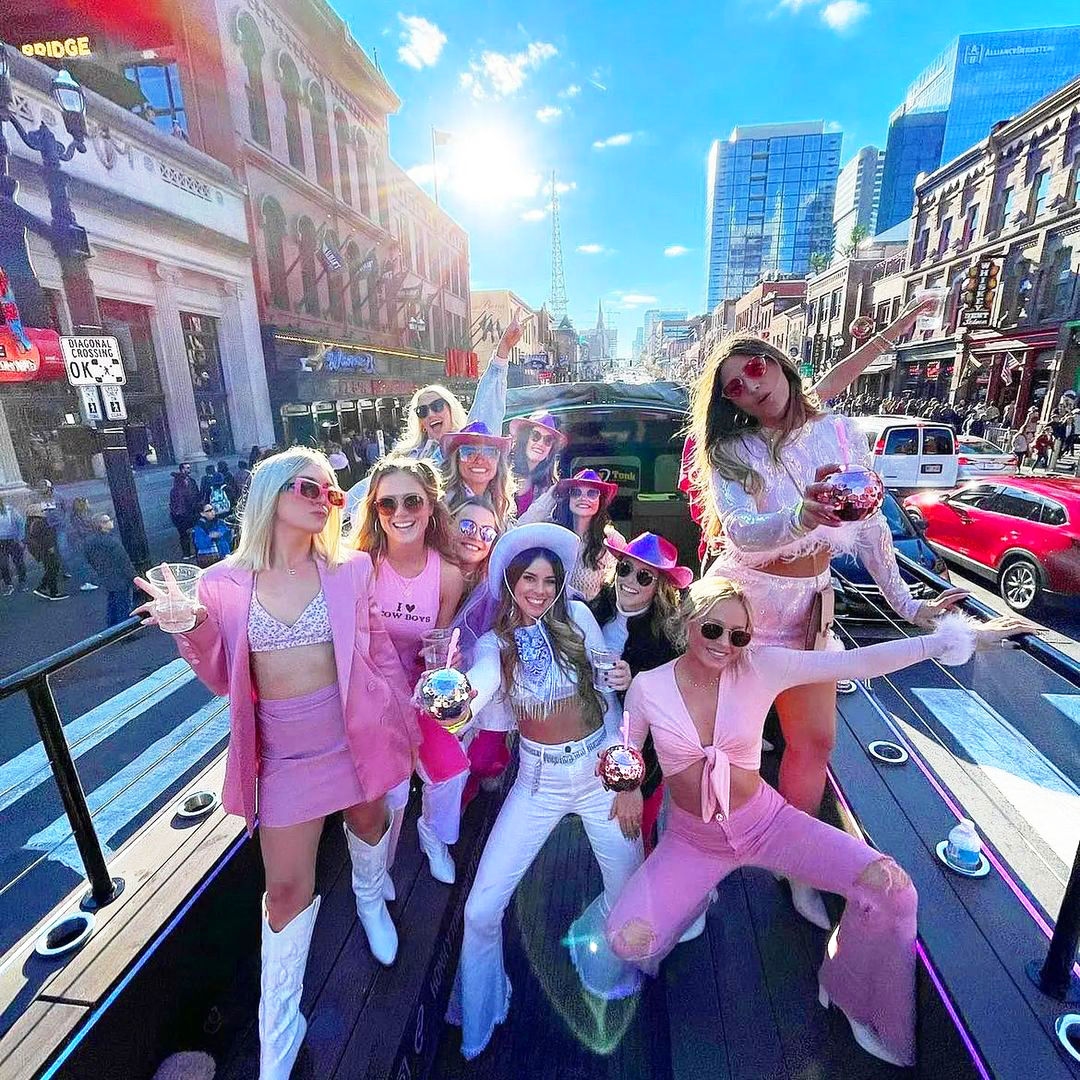 Group of women all wearing pink clothes and some with pink cowboy hats, holding water cups and disco ball glasses on top of a party bus with the city of Nashville in the background
